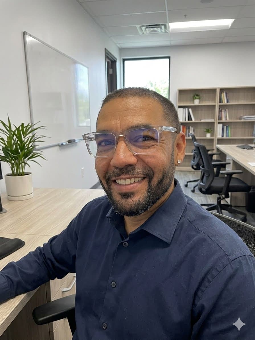 A bearded man in clear glasses and a blue shirt smiles at an office desk.